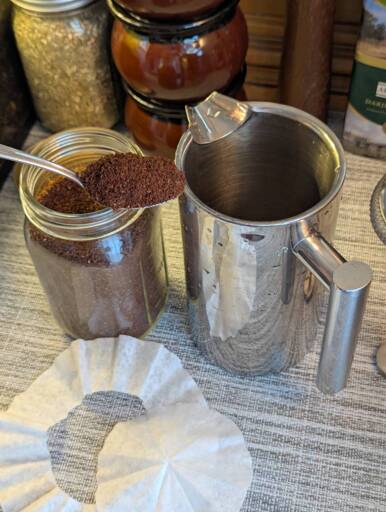 Press coffee maker with a circle of paper filter on the side. A full teaspoon of ground coffee is being held in front of it